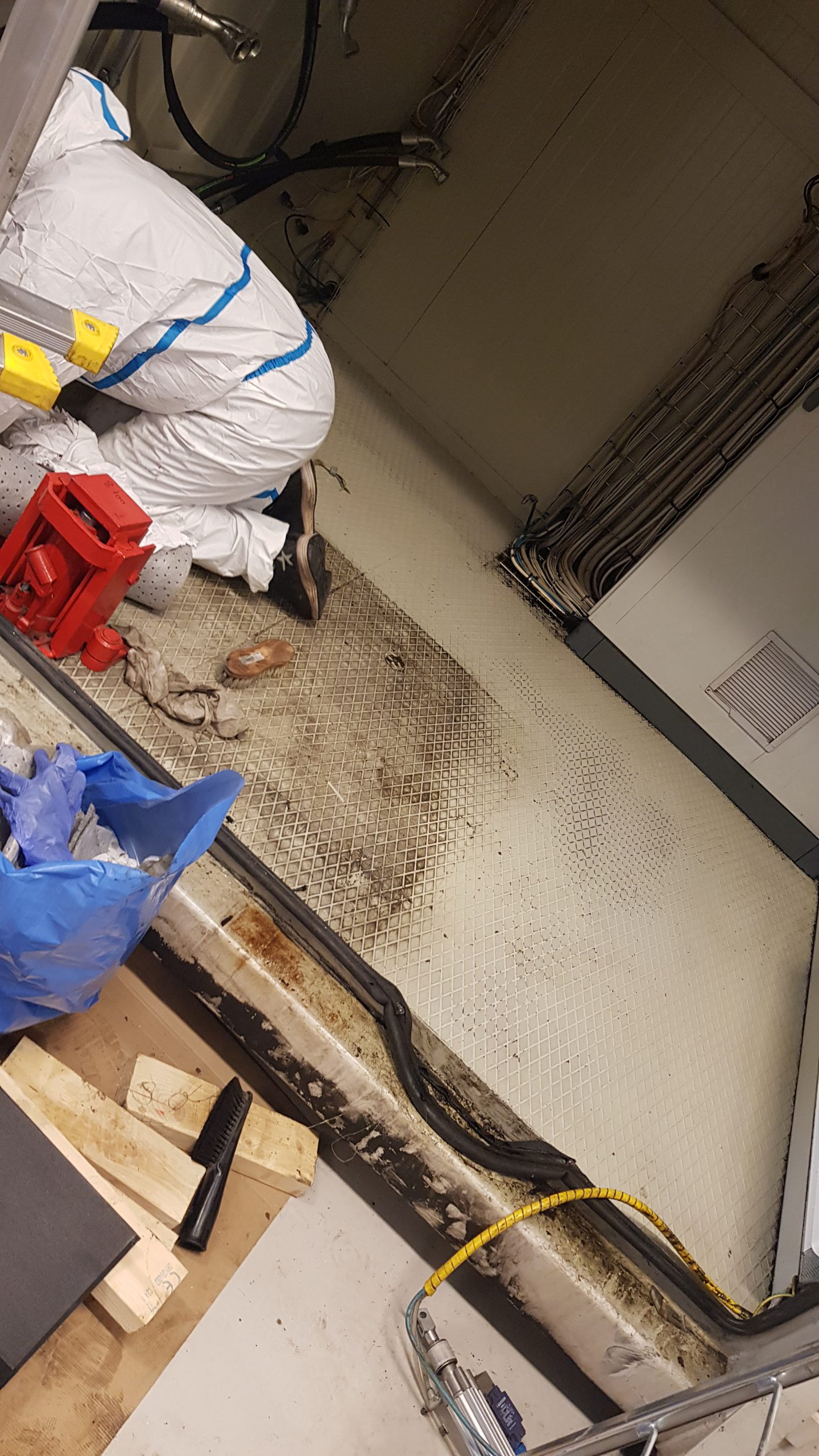 En person i skyddsdräkt och med arbetshandskar är böjd över och arbetar på golvet i ett rum med teknisk utrustning. Rummet har många kablar längs väggen och golvet är täckt av små, vita plattor. Det finns skräp och arbetsverktyg, som en hammare och träblock, utspridda på golvet. Situationen antyder en renovering eller reparation av golvet eller underliggande installationer.
