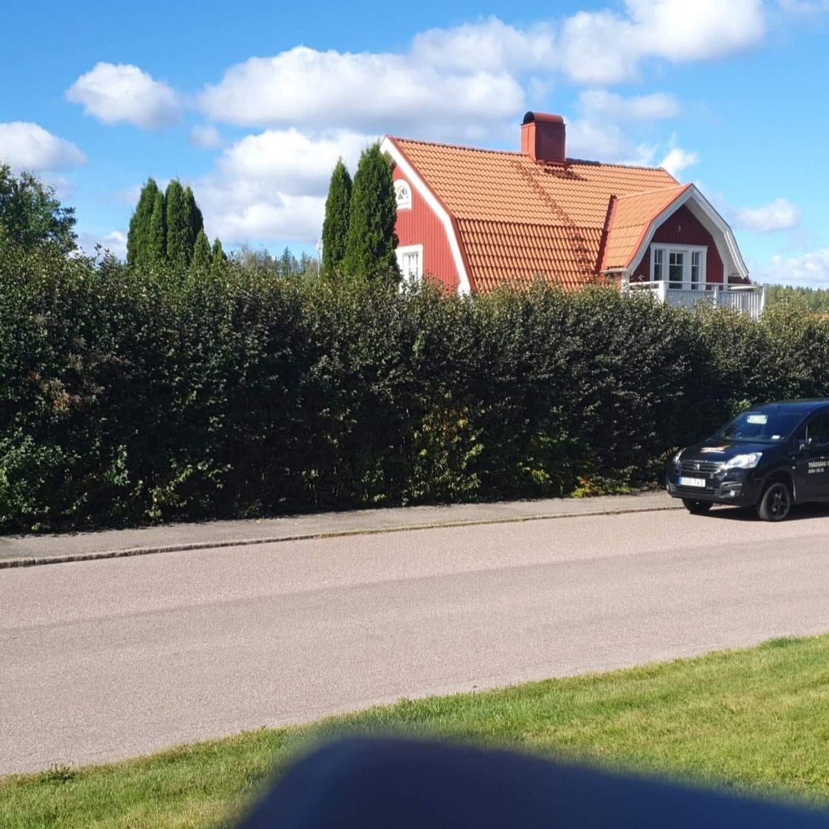 Bilden visar en idyllisk svensk villa med röda tegelväggar och ett mörkt tak, skyddad av en tät häck. Framför huset syns en arbetsbil med släp och en klarblå himmel ovanför, vilket antyder en solig och klar dag.